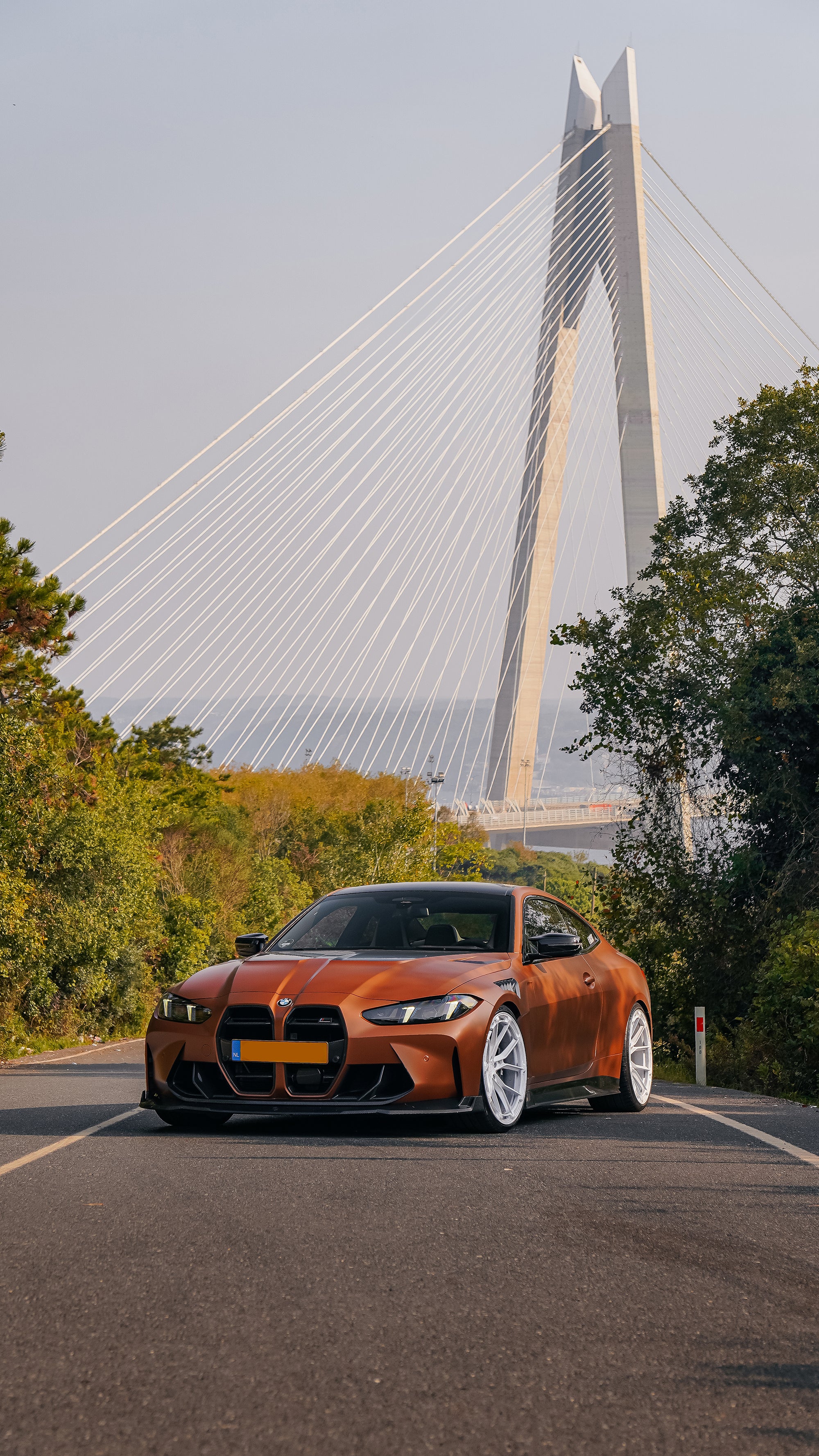Bronze BMW G82 M4 featuring a REVOZPORT carbon fiber body kit with front lip, vented hood, side skirts, and white forged wheels parked on a scenic road near a bridge.