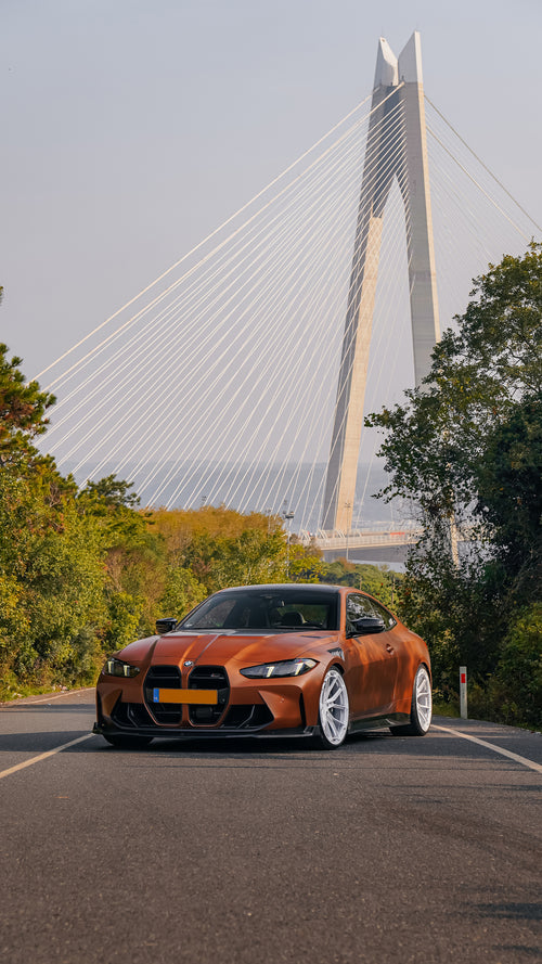 Bronze BMW G82 M4 featuring a REVOZPORT carbon fiber body kit with front lip, vented hood, side skirts, and white forged wheels parked on a scenic road near a bridge.