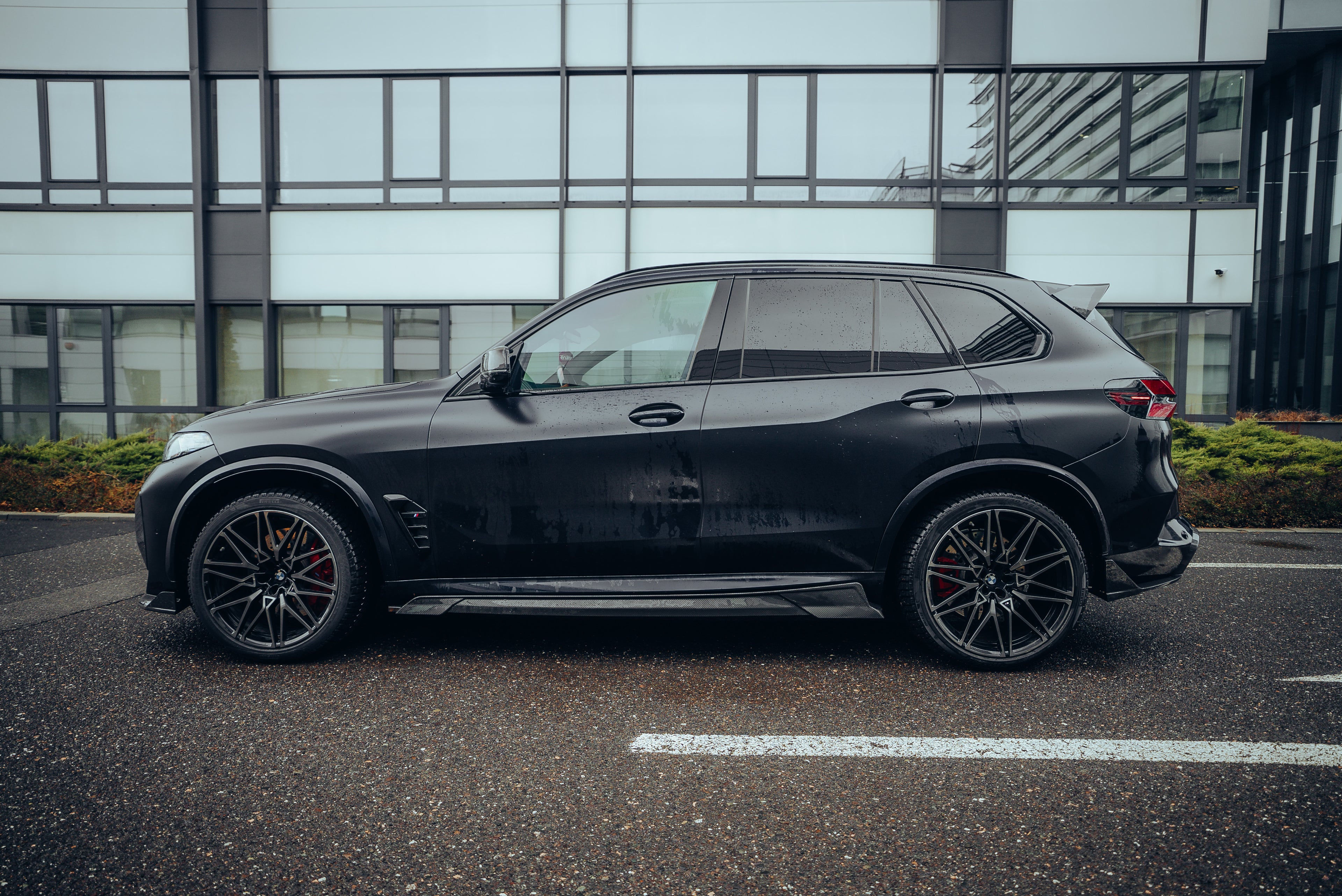 Black BMW X5M featuring a REVOZPORT carbon fiber body kit with aggressive front lip, side skirts, enlarged front air intakes, and sporty aero styling parked in a modern urban business district.