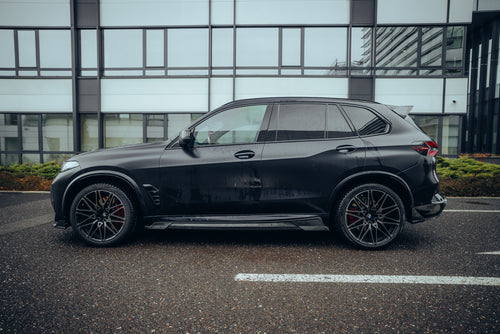 Black BMW X5M featuring a REVOZPORT carbon fiber body kit with aggressive front lip, side skirts, enlarged front air intakes, and sporty aero styling parked in a modern urban business district.