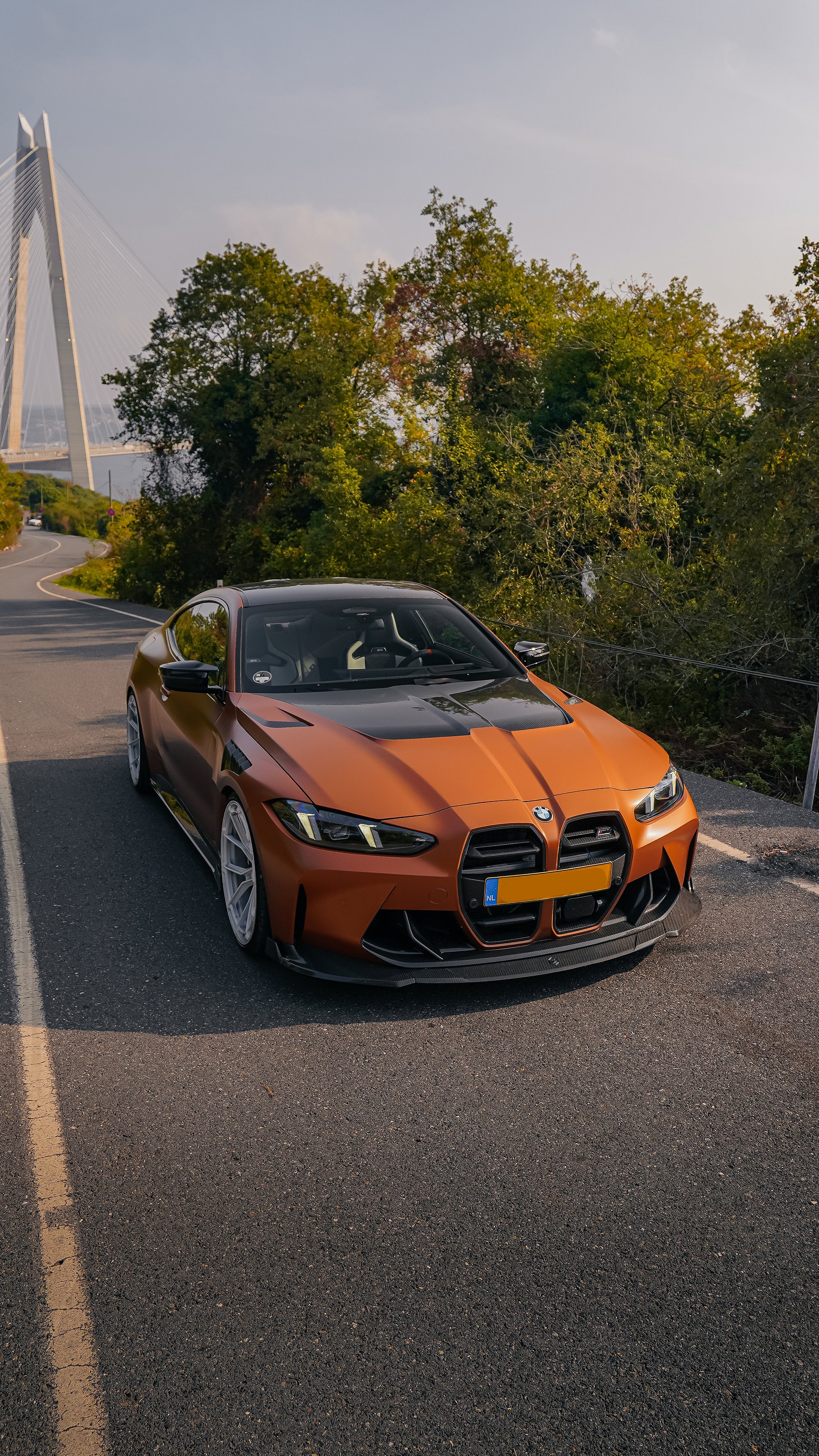 Bronze BMW G82 M4 featuring a REVOZPORT carbon fiber body kit with front lip, vented hood, side skirts, and white forged wheels parked on a scenic road near a bridge.