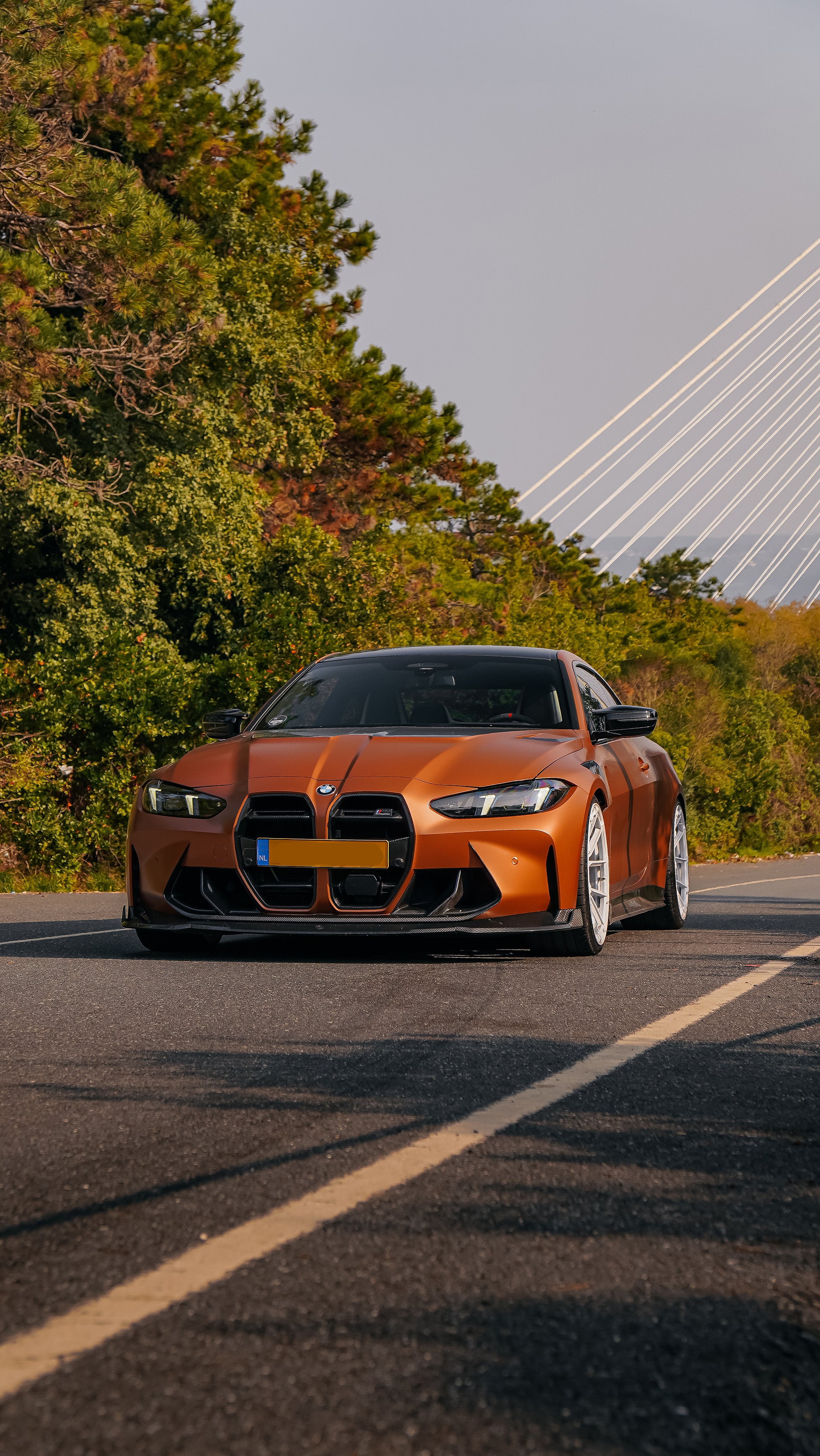 Bronze BMW G82 M4 featuring a REVOZPORT carbon fiber body kit with front lip, vented hood, side skirts, and white forged wheels parked on a scenic road near a bridge.