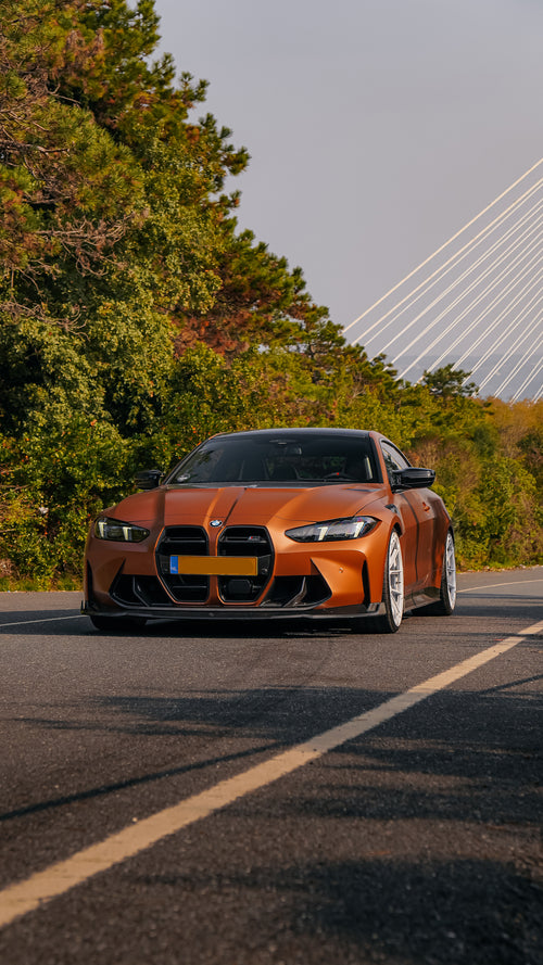 Bronze BMW G82 M4 featuring a REVOZPORT carbon fiber body kit with front lip, vented hood, side skirts, and white forged wheels parked on a scenic road near a bridge.