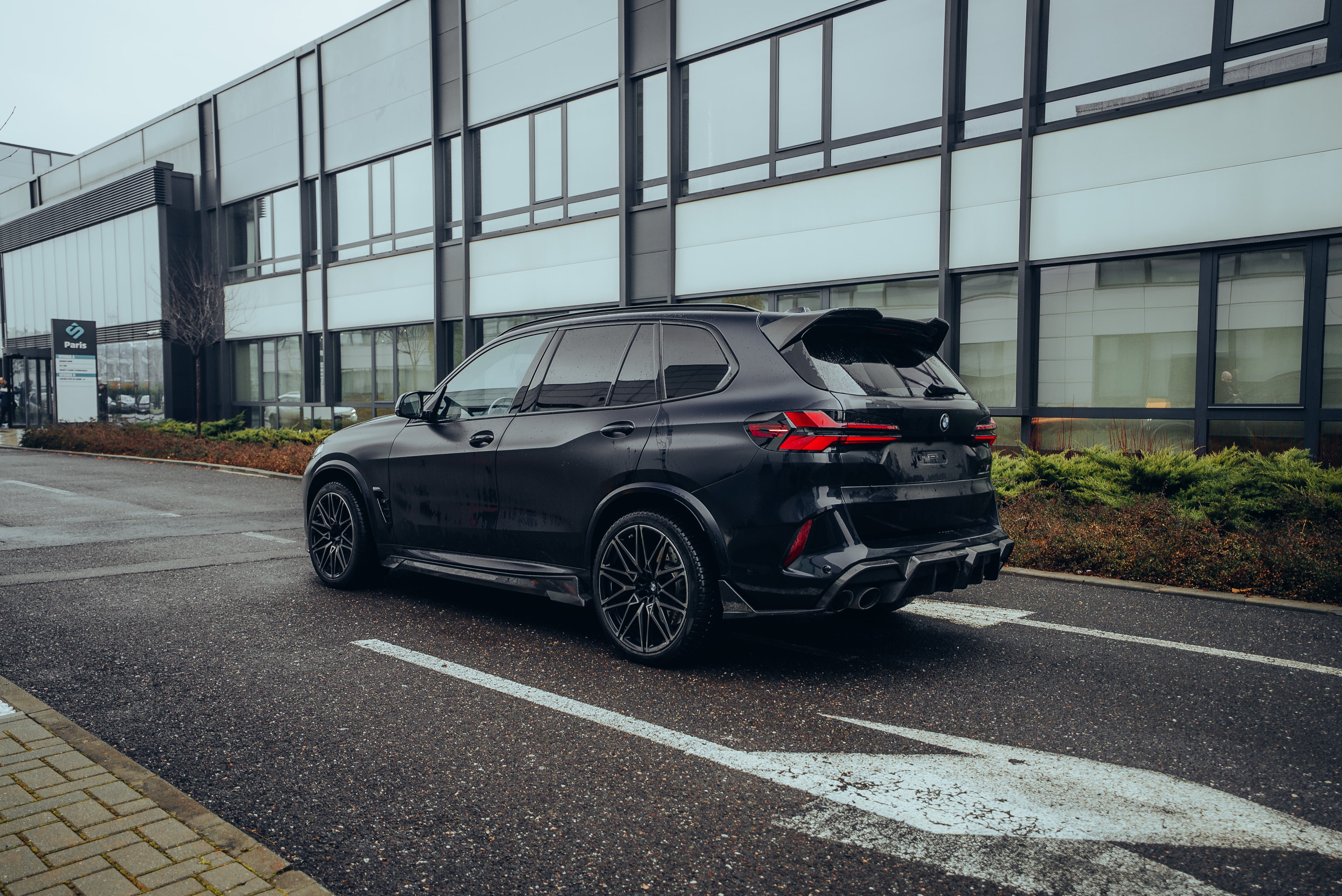 Black BMW X5M featuring a REVOZPORT carbon fiber body kit with aggressive front lip, side skirts, enlarged front air intakes, and sporty aero styling parked in a modern urban business district.