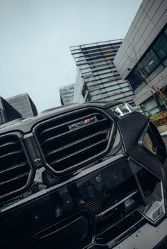Black BMW X5M featuring a REVOZPORT carbon fiber body kit with aggressive front lip, side skirts, enlarged front air intakes, and sporty aero styling parked in a modern urban business district.