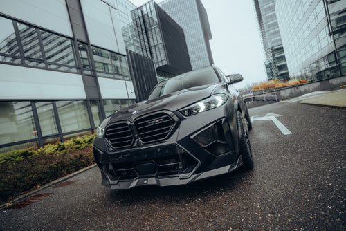 Black BMW X5M featuring a REVOZPORT carbon fiber body kit with aggressive front lip, side skirts, enlarged front air intakes, and sporty aero styling parked in a modern urban business district.