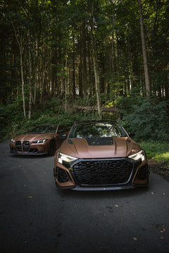 Bronze Audi RS3 featuring a REVOZPORT carbon fiber body kit with aggressive front lip, honeycomb grille, vented hood, and wide aero styling, parked beside a bronze BMW G82 M4 on a forest road.