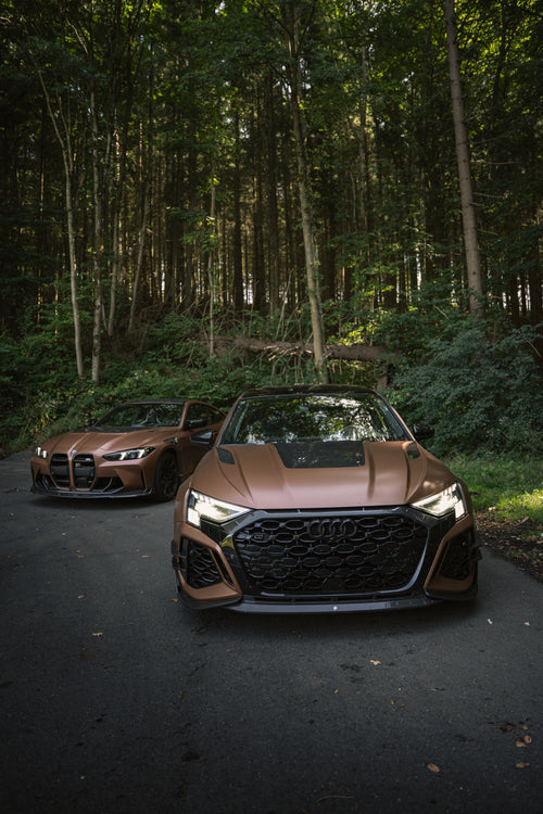 Bronze Audi RS3 featuring a REVOZPORT carbon fiber body kit with aggressive front lip, honeycomb grille, vented hood, and wide aero styling, parked beside a bronze BMW G82 M4 on a forest road.