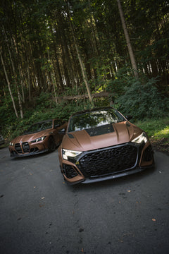 Bronze Audi RS3 featuring a REVOZPORT carbon fiber body kit with aggressive front lip, honeycomb grille, vented hood, and wide aero styling, parked beside a bronze BMW G82 M4 on a forest road.