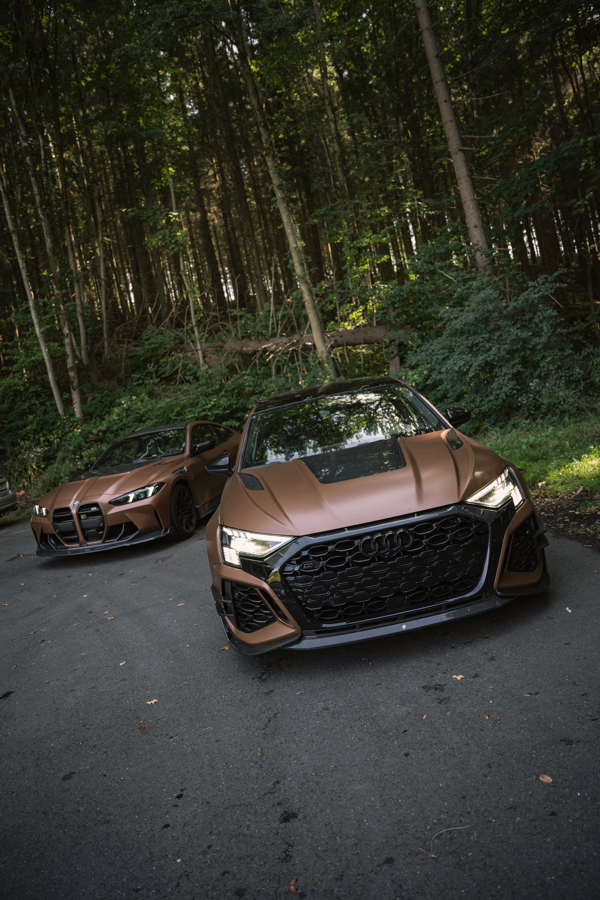 Bronze Audi RS3 featuring a REVOZPORT carbon fiber body kit with aggressive front lip, honeycomb grille, vented hood, and wide aero styling, parked beside a bronze BMW G82 M4 on a forest road.