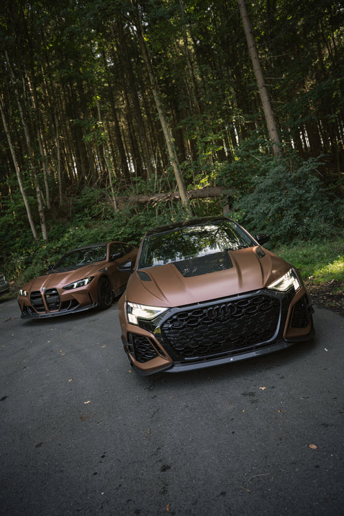 Bronze Audi RS3 featuring a REVOZPORT carbon fiber body kit with aggressive front lip, honeycomb grille, vented hood, and wide aero styling, parked beside a bronze BMW G82 M4 on a forest road.