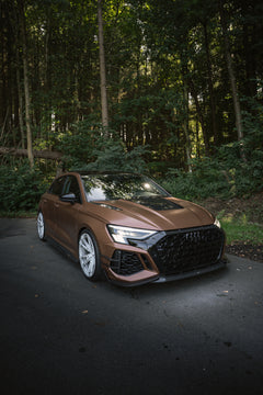 Bronze Audi RS3 featuring a REVOZPORT carbon fiber body kit with aggressive front lip, honeycomb grille, vented hood, and wide aero styling, parked beside a bronze BMW G82 M4 on a forest road.