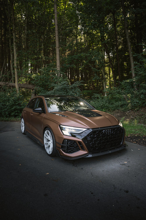 Bronze Audi RS3 featuring a REVOZPORT carbon fiber body kit with aggressive front lip, honeycomb grille, vented hood, and wide aero styling, parked beside a bronze BMW G82 M4 on a forest road.