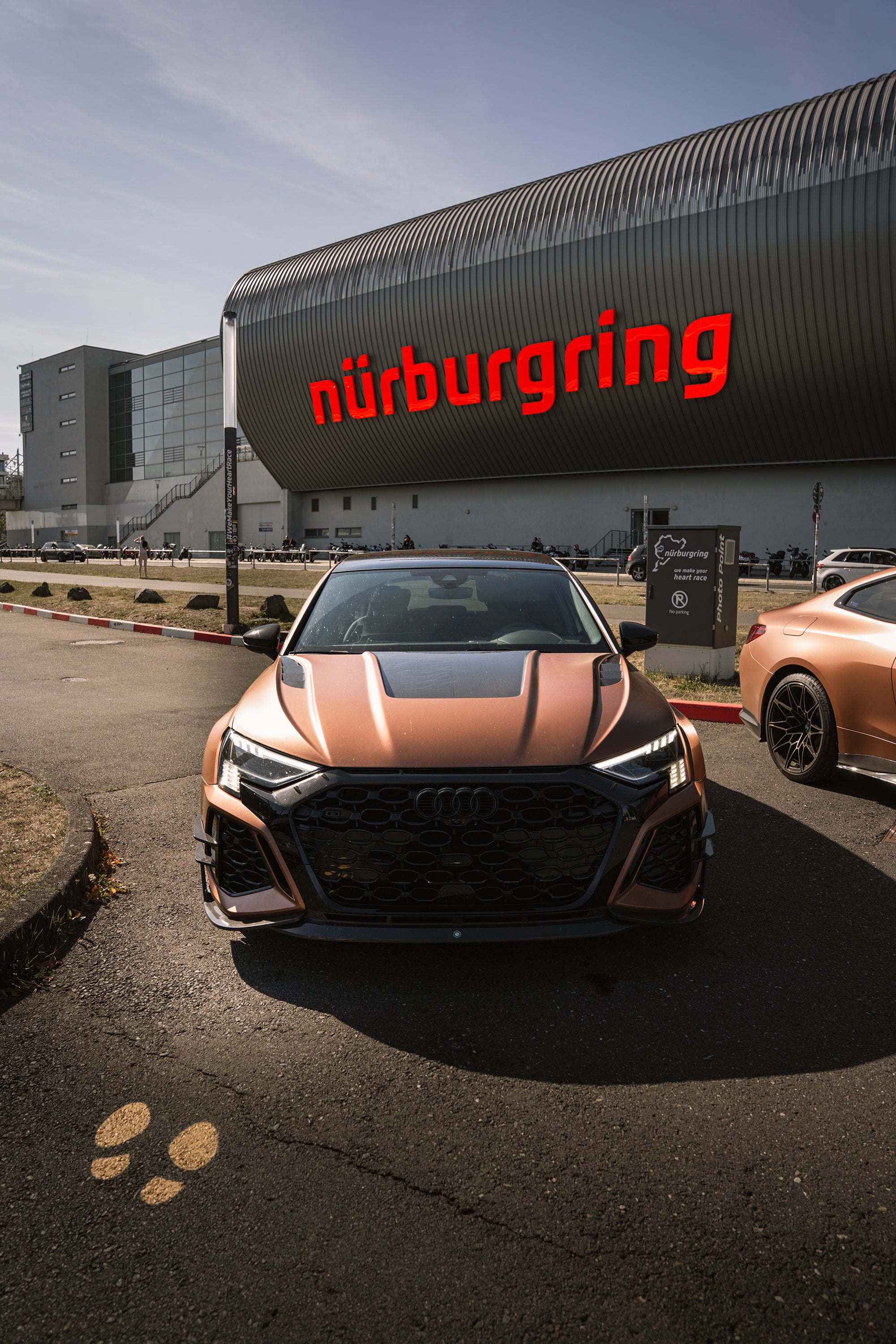Bronze Audi RS3 featuring a REVOZPORT carbon fiber body kit with aggressive front lip, honeycomb grille, vented hood, and wide aero styling, parked beside a bronze BMW G82 M4 on a forest road.