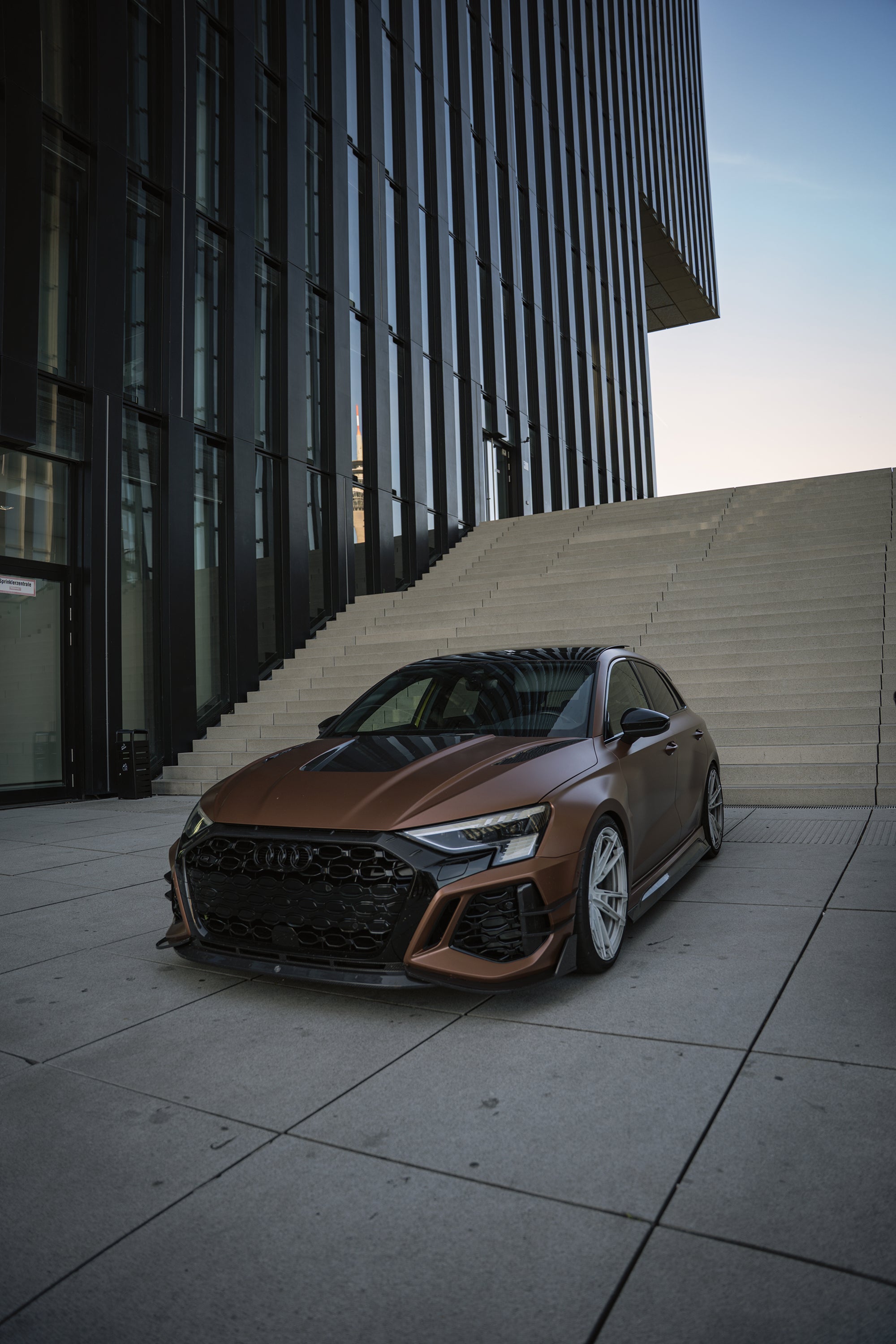 Bronze Audi RS3 featuring a REVOZPORT carbon fiber body kit with aggressive front lip, honeycomb grille, vented hood, and wide aero styling, parked beside a bronze BMW G82 M4 on a forest road.