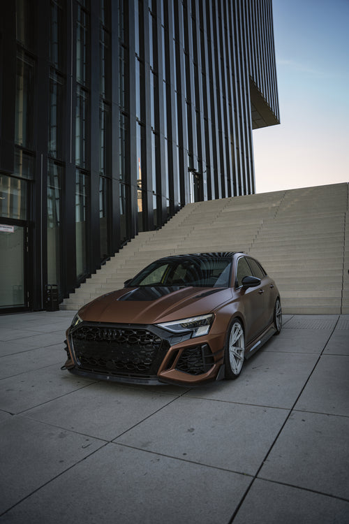 Bronze Audi RS3 featuring a REVOZPORT carbon fiber body kit with aggressive front lip, honeycomb grille, vented hood, and wide aero styling, parked beside a bronze BMW G82 M4 on a forest road.