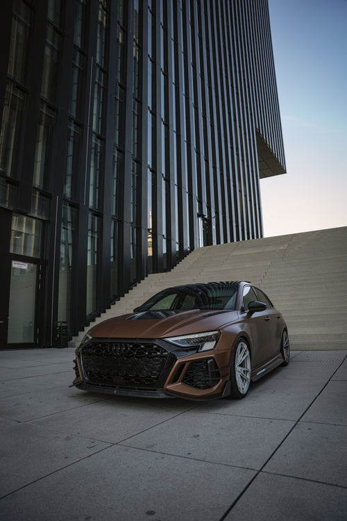 Bronze Audi RS3 featuring a REVOZPORT carbon fiber body kit with aggressive front lip, honeycomb grille, vented hood, and wide aero styling, parked beside a bronze BMW G82 M4 on a forest road.