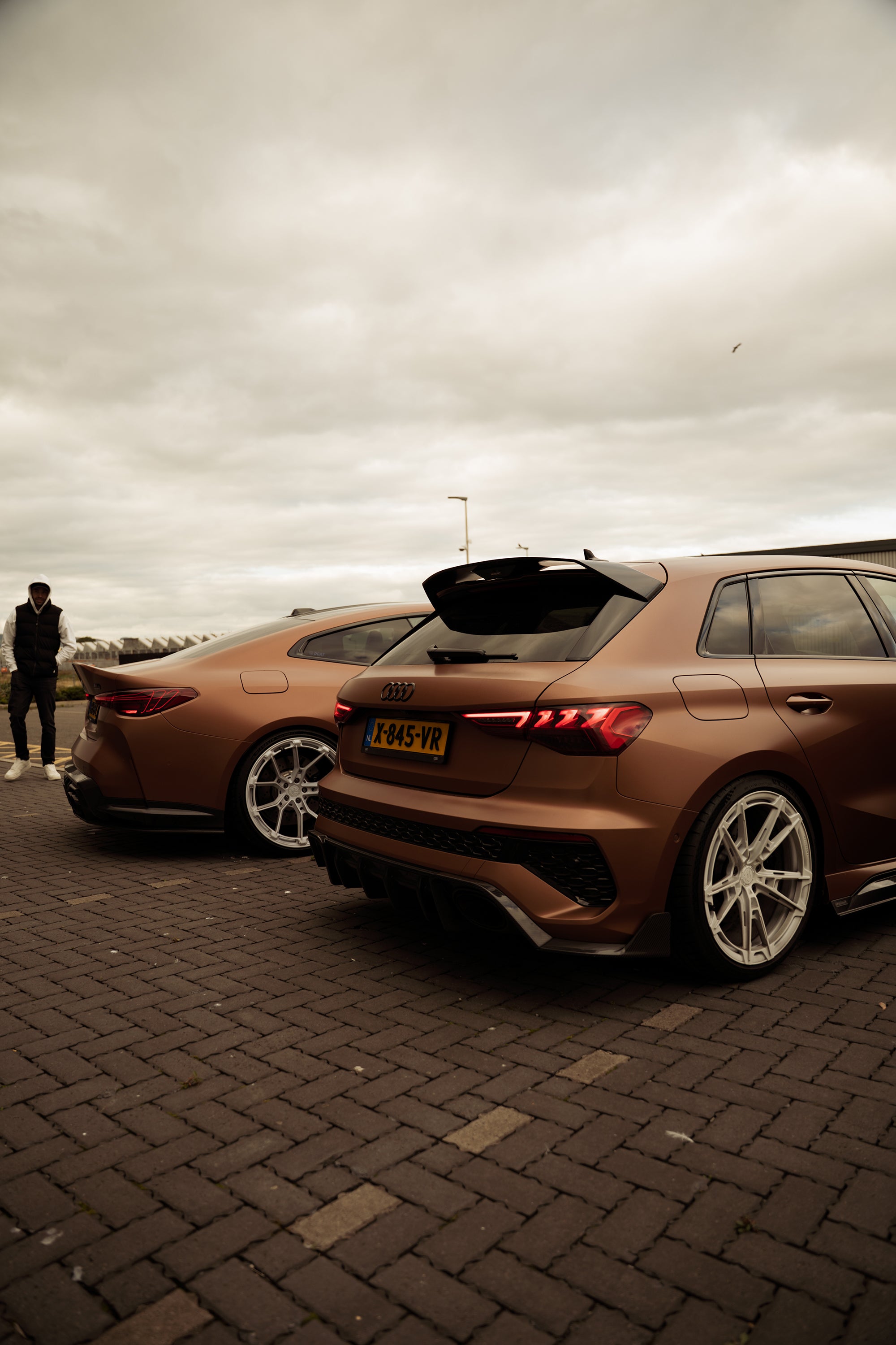 Bronze Audi RS4 Avant featuring a REVOZPORT carbon fiber body kit with front lip and side skirts, paired with silver multi-spoke forged wheels, parked in a modern urban setting.