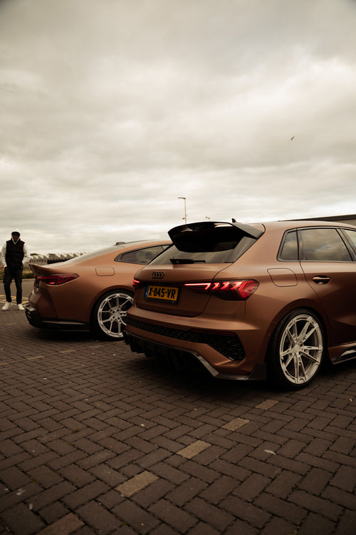 Bronze Audi RS4 Avant featuring a REVOZPORT carbon fiber body kit with front lip and side skirts, paired with silver multi-spoke forged wheels, parked in a modern urban setting.