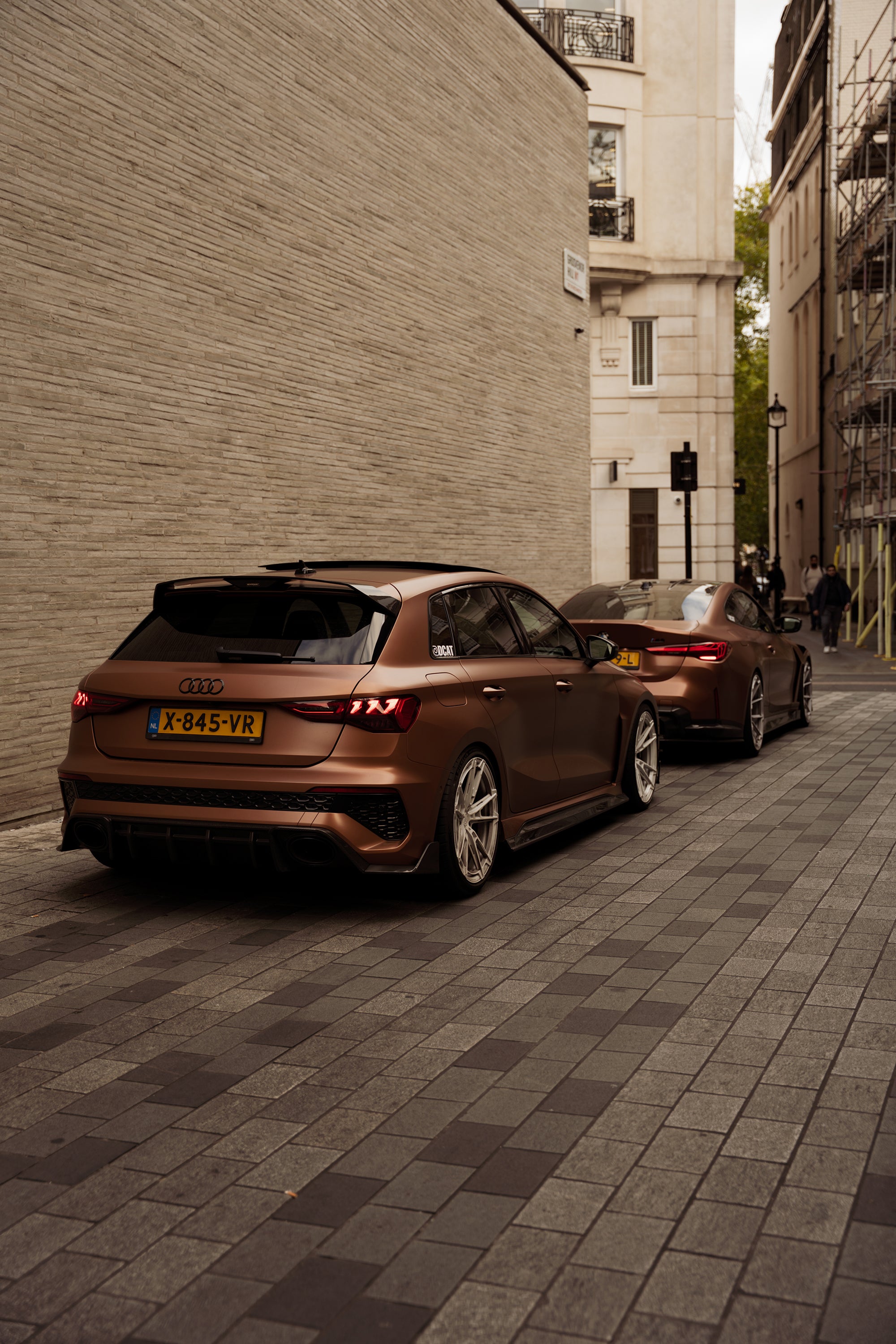 Bronze Audi RS4 Avant featuring a REVOZPORT carbon fiber body kit with front lip and side skirts, paired with silver multi-spoke forged wheels, parked in a modern urban setting.