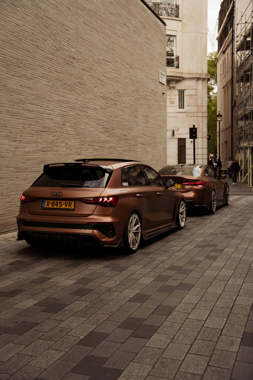 Bronze Audi RS4 Avant featuring a REVOZPORT carbon fiber body kit with front lip and side skirts, paired with silver multi-spoke forged wheels, parked in a modern urban setting.