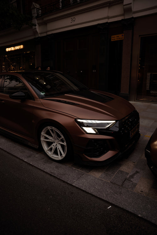 Bronze Audi RS4 Avant featuring a REVOZPORT carbon fiber body kit with front lip and side skirts, paired with silver multi-spoke forged wheels, parked in a modern urban setting.