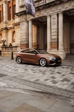 Bronze Audi RS4 Avant featuring a REVOZPORT carbon fiber body kit with front lip and side skirts, paired with silver multi-spoke forged wheels, parked in a modern urban setting.