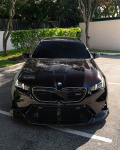 BMW touring car with carbon fiber body kit parked in a leafy outdoor lot, highlighting front lip and lowered stance.