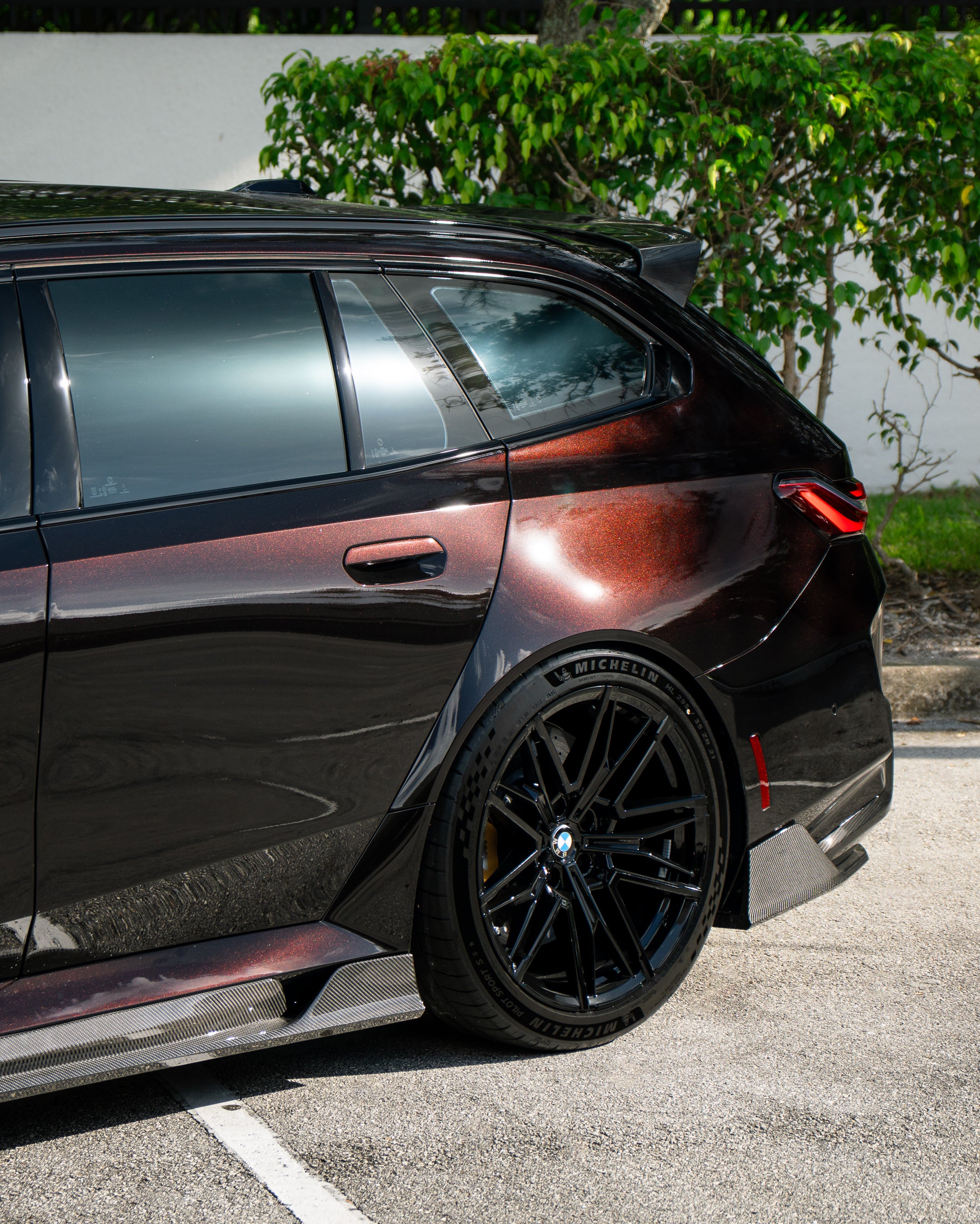 Close-up of a BMW rear wheel with carbon fiber side skirt and diffuser detail, highlighting glossy weave and performance styling.