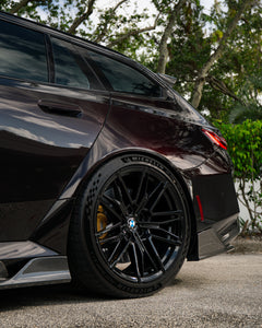 Close-up of a BMW rear wheel with carbon fiber side skirt and diffuser detail, highlighting glossy weave and performance styling.