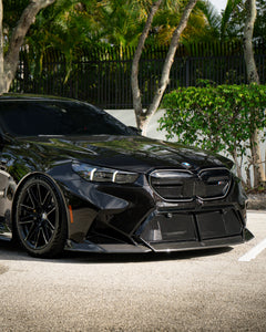 BMW touring car with carbon fiber body kit parked in a leafy outdoor lot, highlighting front lip and lowered stance.