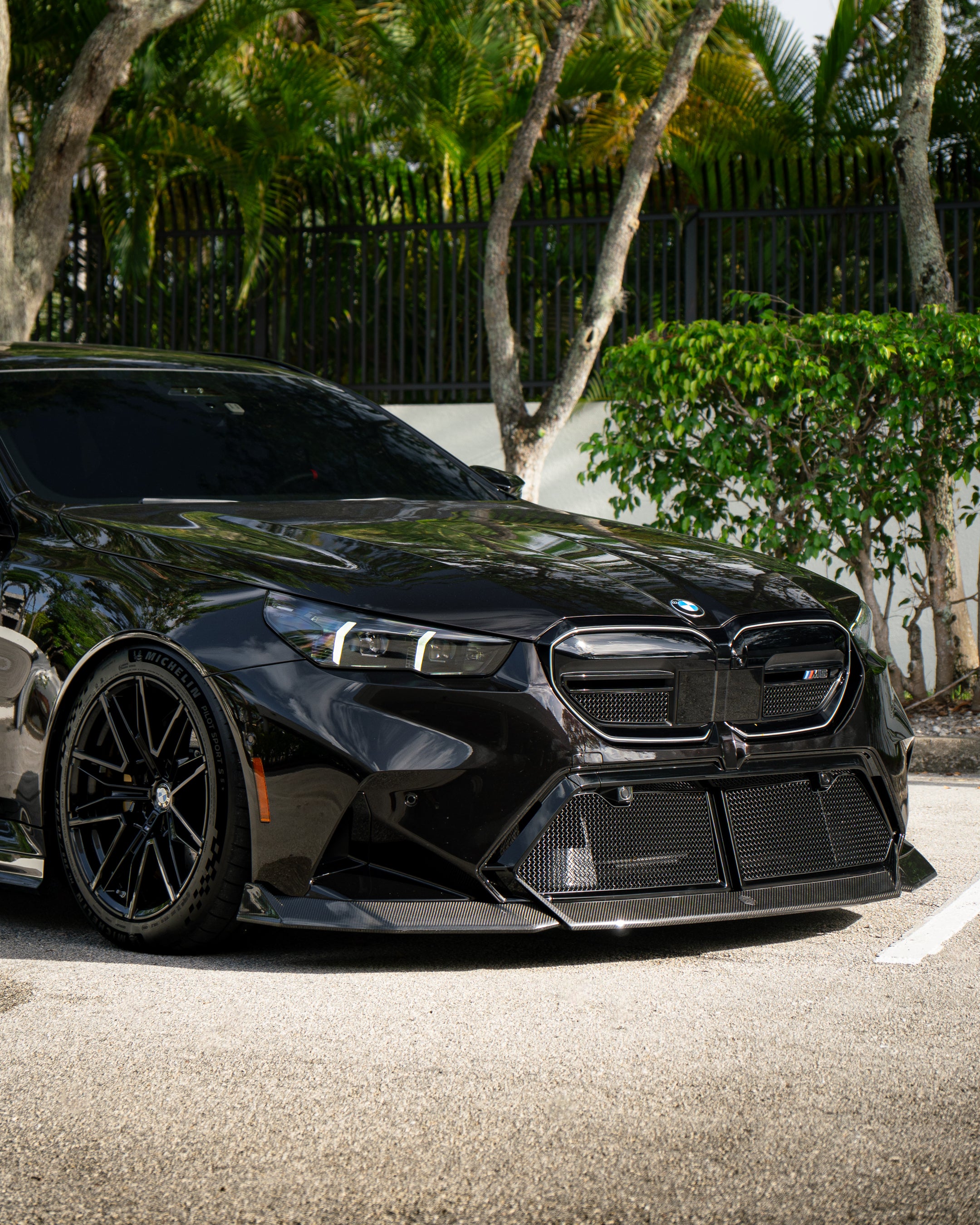 BMW touring car with carbon fiber body kit parked in a leafy outdoor lot, highlighting front lip and lowered stance.
