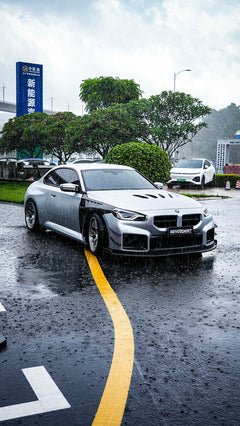 White BMW G82 M4 with carbon fiber hood, vented fenders, side skirts, and oversized GT wing driving through heavy rain on a wet urban road.