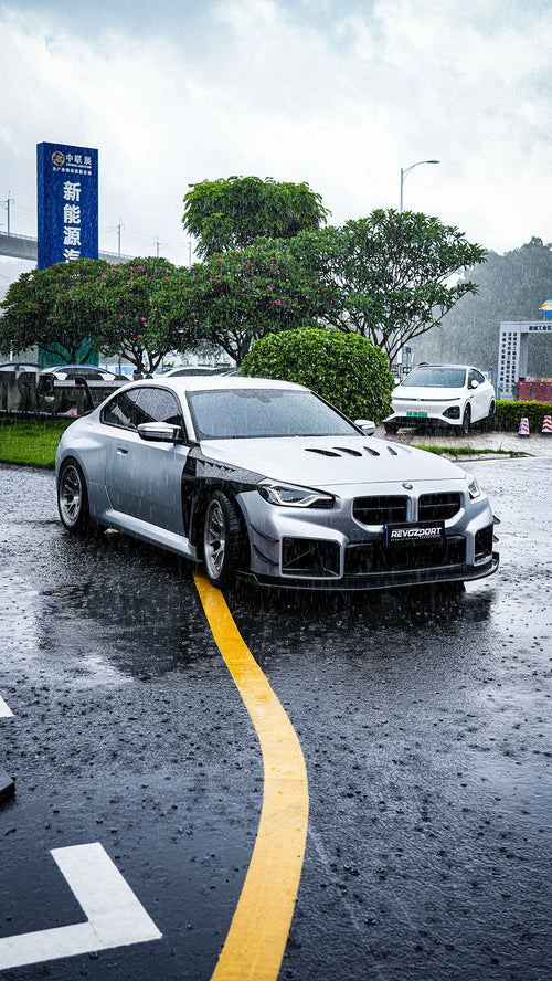 White BMW G82 M4 with carbon fiber hood, vented fenders, side skirts, and oversized GT wing driving through heavy rain on a wet urban road.
