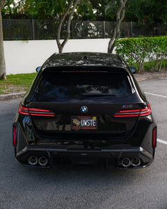 Rear three-quarter view of a BMW touring car with carbon fiber aero kit, rear diffuser, roof spoiler, and quad exhausts parked in a leafy outdoor setting.