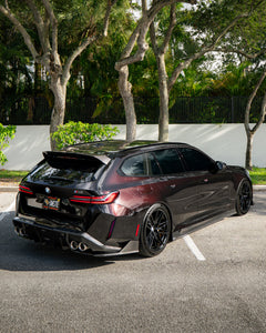 Rear three-quarter view of a BMW touring car with carbon fiber aero kit, rear diffuser, roof spoiler, and quad exhausts parked in a leafy outdoor setting.