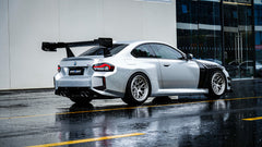 White BMW G82 M4 with carbon fiber hood, vented fenders, side skirts, and oversized GT wing driving through heavy rain on a wet urban road.