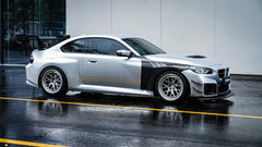 White BMW G82 M4 with carbon fiber hood, vented fenders, side skirts, and oversized GT wing driving through heavy rain on a wet urban road.