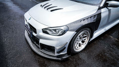 Close-up of a BMW with carbon fiber widebody kit, front splitter, vented hood, and white wheels on a rainy street.