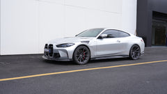 Silver BMW G82 M4 featuring a REVOZPORT carbon fiber body kit with front lip, side skirts, vented fenders, and aggressive aero styling parked outside a modern building.