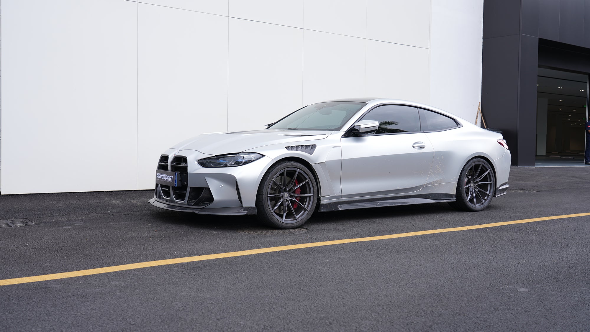 Silver BMW G82 M4 featuring a REVOZPORT carbon fiber body kit with front lip, side skirts, vented fenders, and aggressive aero styling parked outside a modern building.