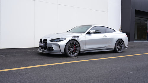 Silver BMW G82 M4 featuring a REVOZPORT carbon fiber body kit with front lip, side skirts, vented fenders, and aggressive aero styling parked outside a modern building.