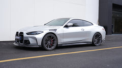 Silver BMW G82 M4 featuring a REVOZPORT carbon fiber body kit with front lip, side skirts, vented fenders, and aggressive aero styling parked outside a modern building.
