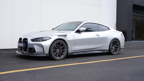 Silver BMW G82 M4 featuring a REVOZPORT carbon fiber body kit with front lip, side skirts, vented fenders, and aggressive aero styling parked outside a modern building.