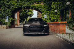 Black Audi with carbon fiber hood and front lip parked under an ivy-covered arch, showcasing aggressive front aero styling.