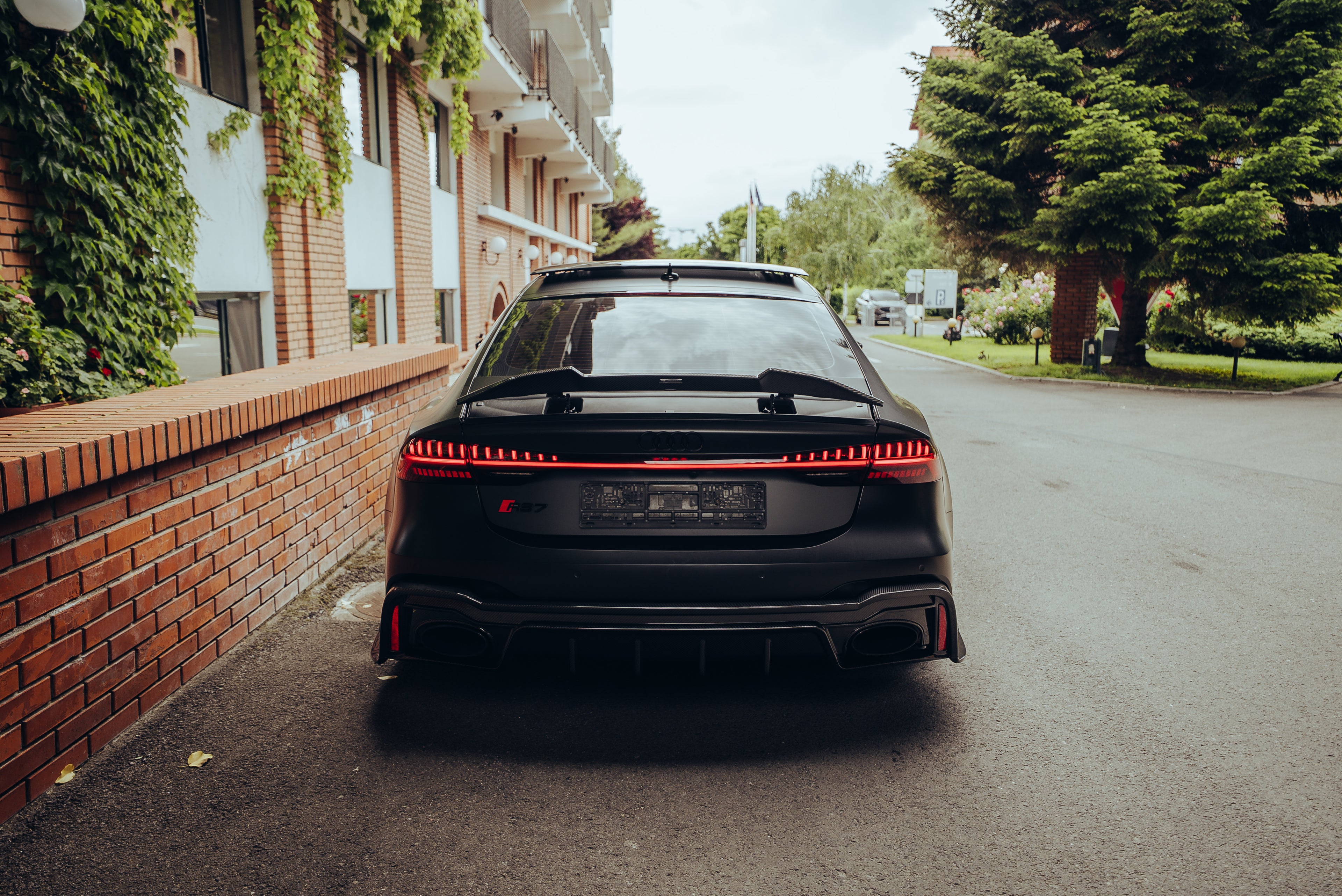 Rear view of a matte black Audi with carbon fiber rear spoiler and diffuser, parked beside ivy-covered brick architecture.