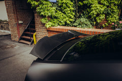 Close-up of a carbon fiber rear spoiler, highlighting glossy weave texture and sporty aero styling in an urban setting.