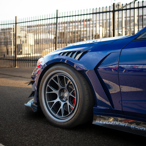 Blue BMW G80 M3 with aggressive carbon fiber aero kit, front splitter, vented fenders, and side skirts parked beside a professional race track during sunset.