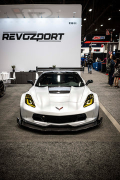 Silver Chevrolet Corvette race car displayed at an indoor automotive exhibition, featuring a wide-body aerodynamic kit, large rear wing, carbon fiber front splitter, and vented hood. The aggressive motorsport styling is showcased under exhibition lighting with REVOZPORT branding in the background.