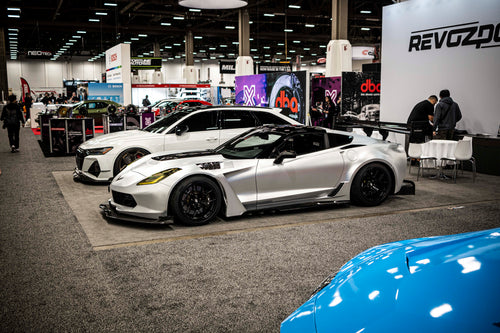 Silver Chevrolet Corvette race car displayed at an indoor automotive exhibition, featuring a wide-body aerodynamic kit, large rear wing, carbon fiber front splitter, and vented hood. The aggressive motorsport styling is showcased under exhibition lighting with REVOZPORT branding in the background.